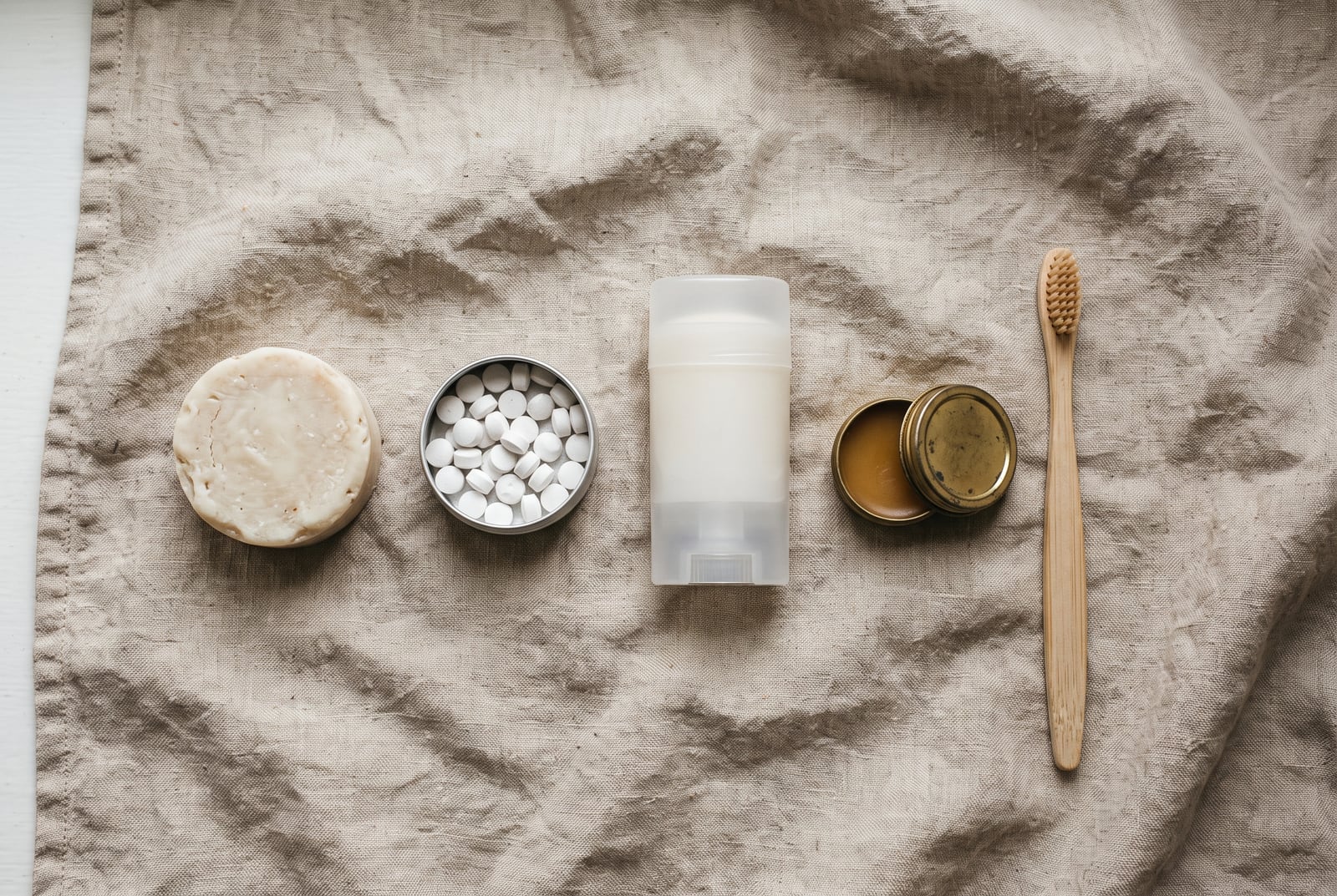 A bar of soap, an open tin of white toothpaste tablets, a plain solid deodorant stick, a small brass tin of balm and a wooden toothbrush laid out in a row on rumpled linen