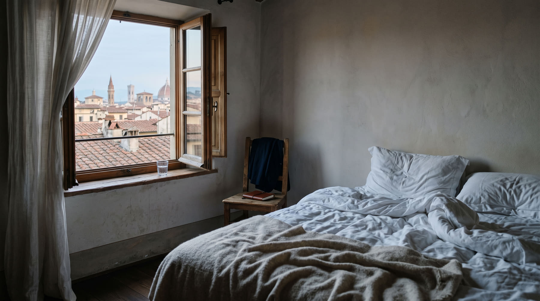 An unmade hotel bed and an open window over terracotta rooftops