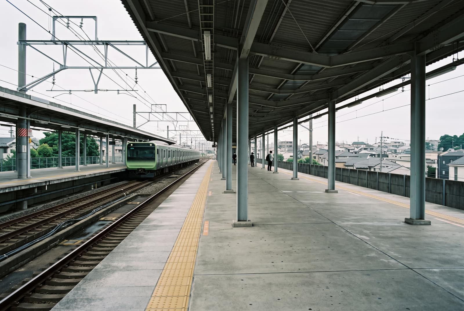A quiet above-ground commuter railway platform in a Tokyo suburb, a train stopped at the far end, a few distant figures